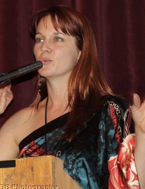 jamie kristen with red hair at podium speaking with hands off-shoulder blouse in teal and coral